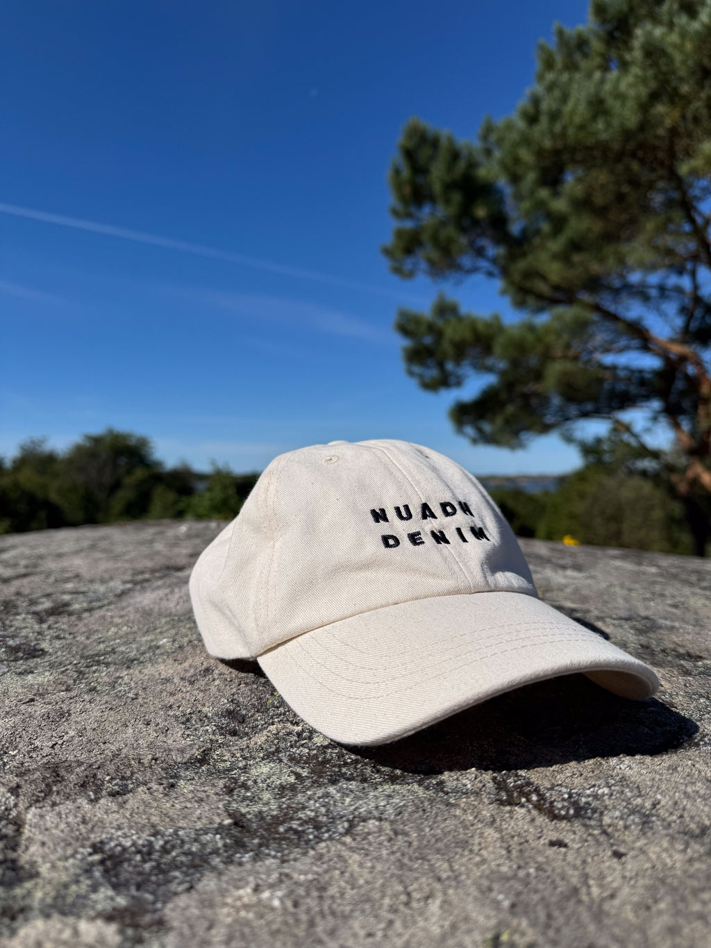 Natural unbleached NUADH DENIM - Sannox bay - Dad cap with embroidered NUADH DENIM logo in navy sitting on a stone wall with trees and blue sky in the background.

NUADH DENIM | Catacol Bay Dad cap | 100% Organic unbleached cotton| Made in Scotland.