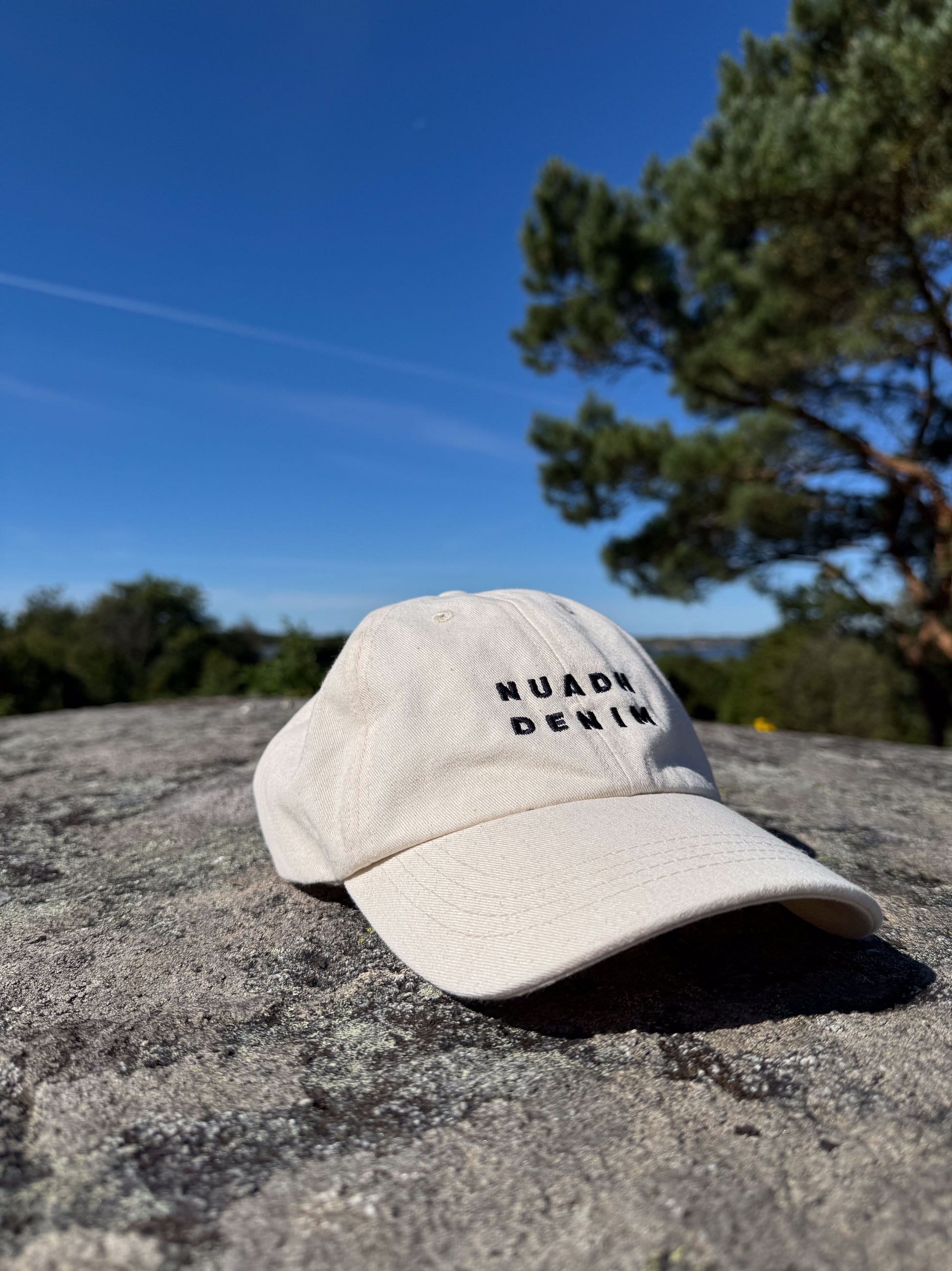 Natural unbleached NUADH DENIM - Sannox bay - Dad cap with embroidered NUADH DENIM logo in navy sitting on a stone wall with trees and blue sky in the background.

NUADH DENIM | Catacol Bay Dad cap | 100% Organic unbleached cotton| Made in Scotland.